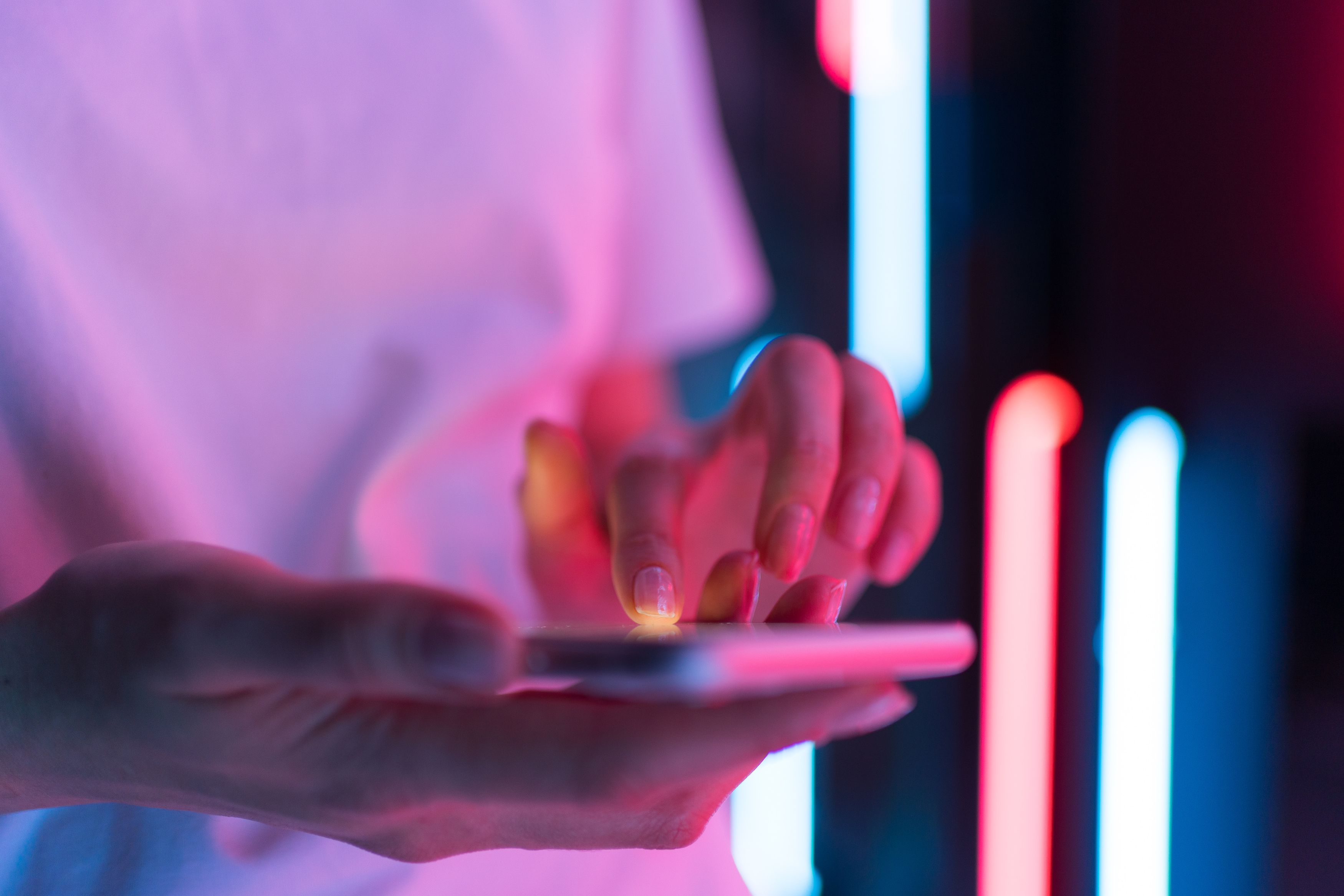 A person is tapping on a smart phone with neon pink and blue lights in the background.