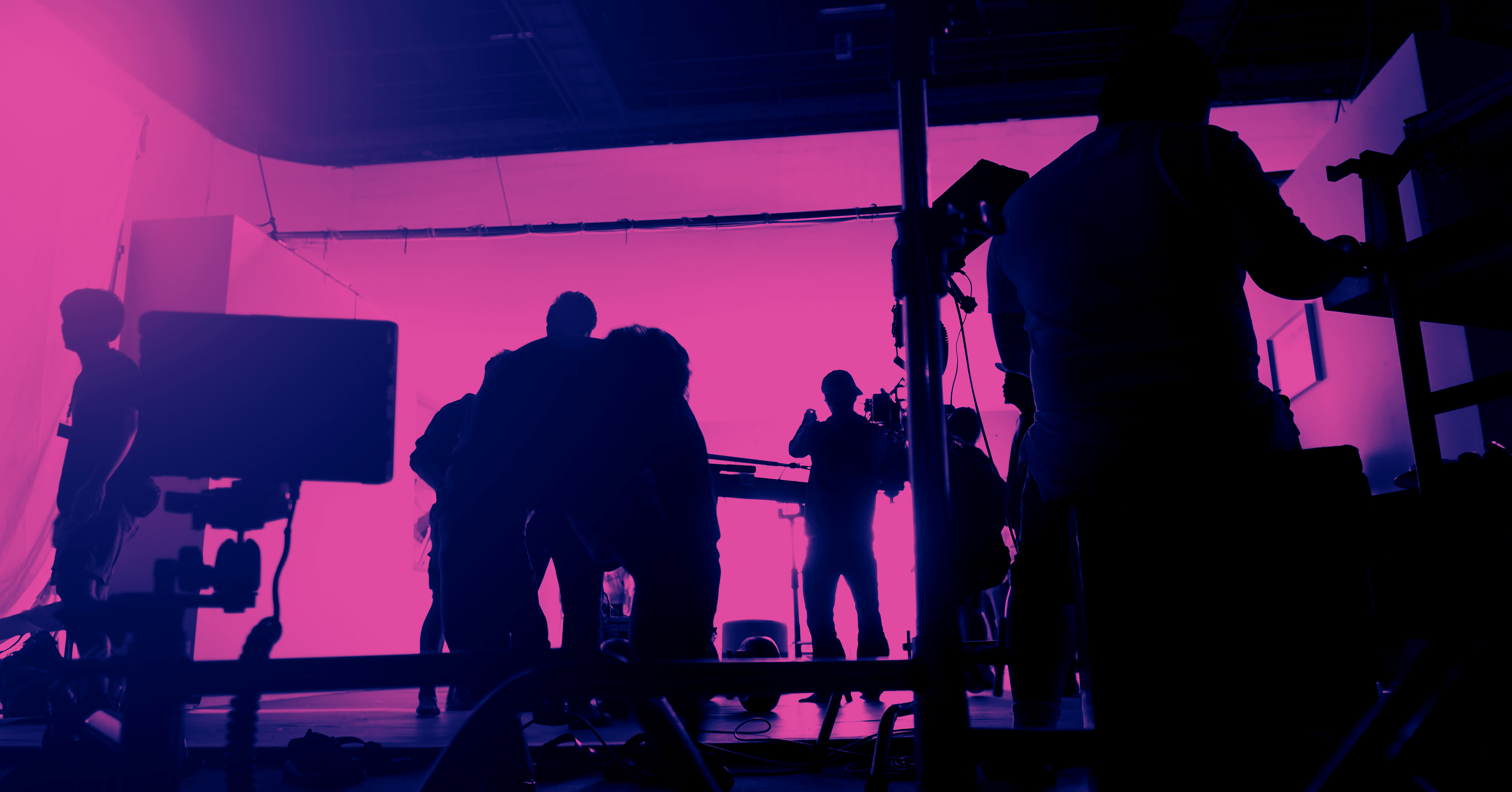 A full visual crew is setting up in front of backdrop at a shoot in a dark room with pink light