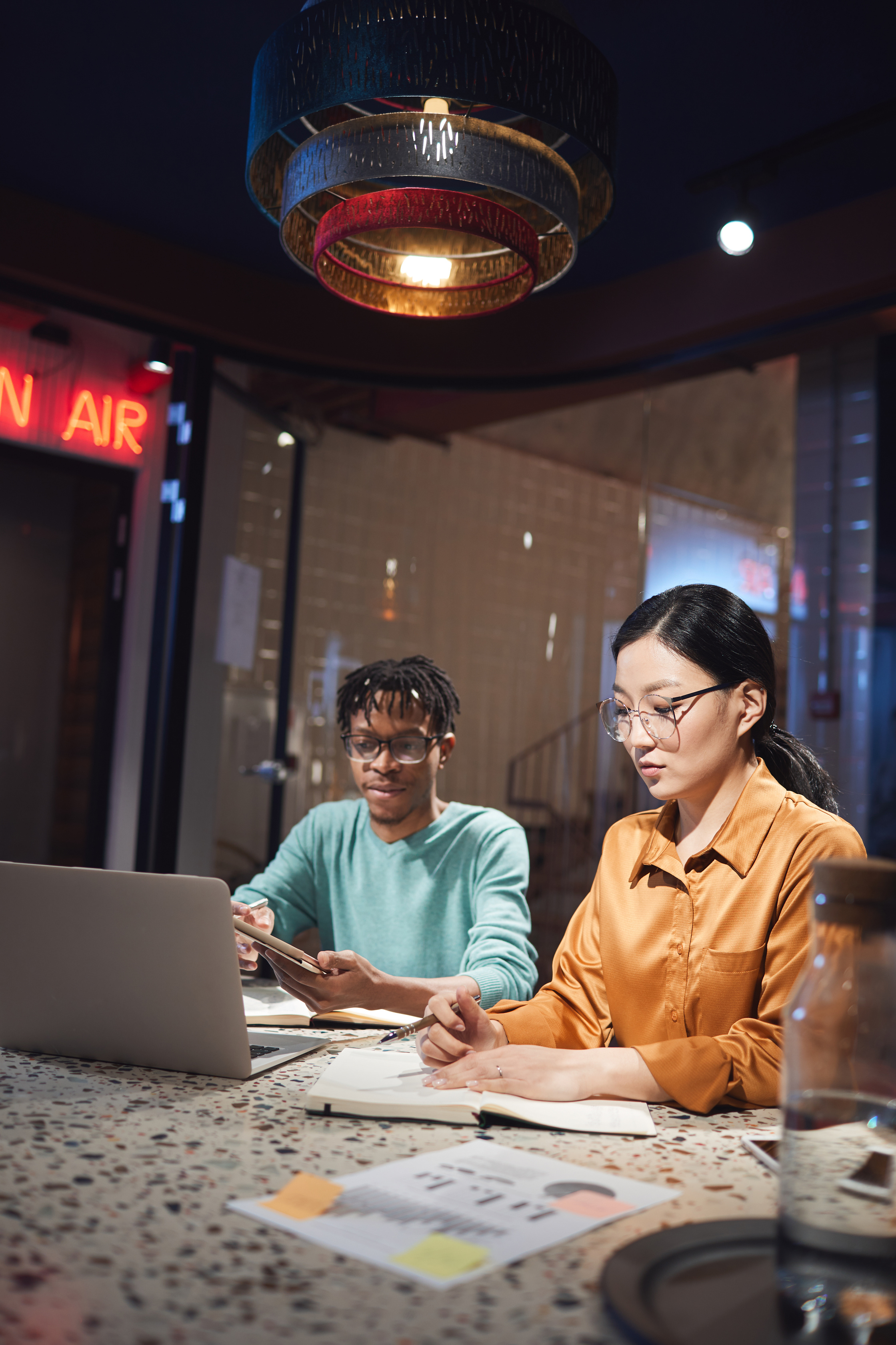 Two people sit together around a desk with a computer and various papers stretched out in front of them. 